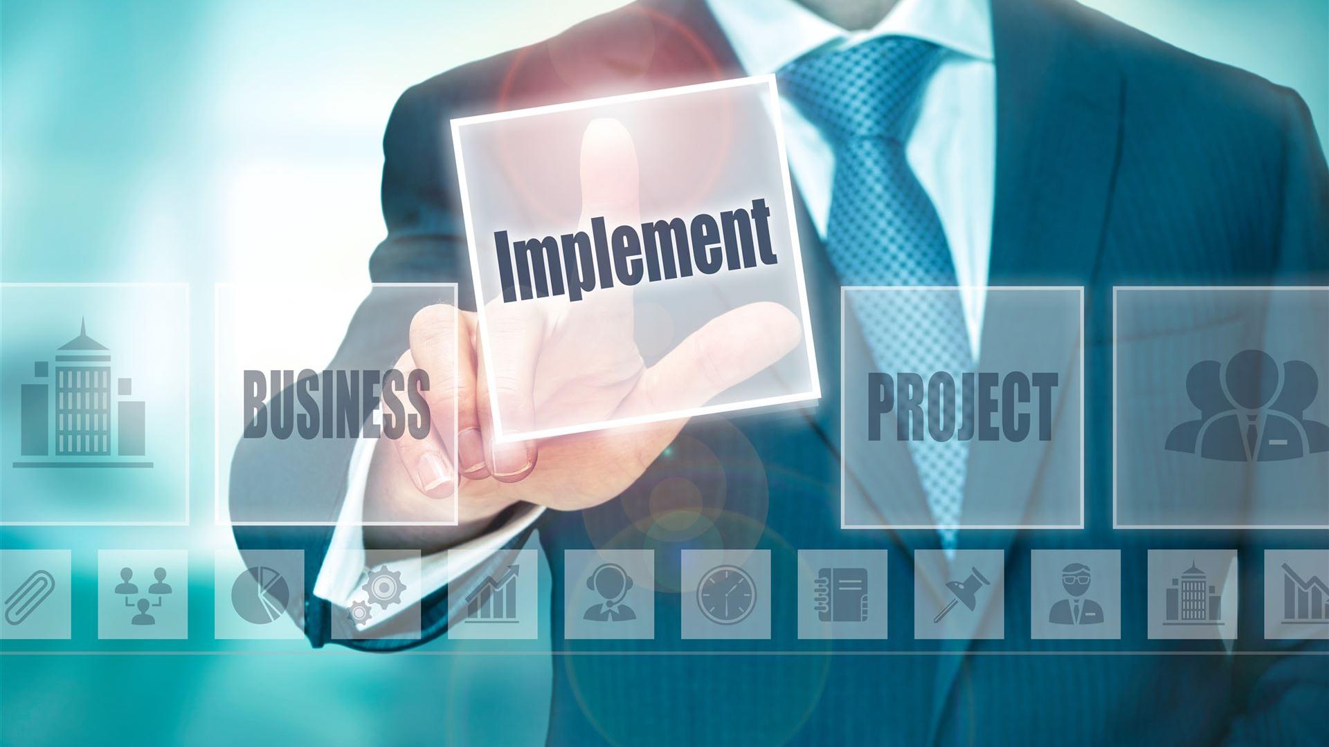 Business-Mann in Anzug tippt auf ein transparentes digitales Interface mit dem Begriff „Implement“ – symbolisiert unsere Umsetzung von Business-Projekten und strategischen Maßnahmen